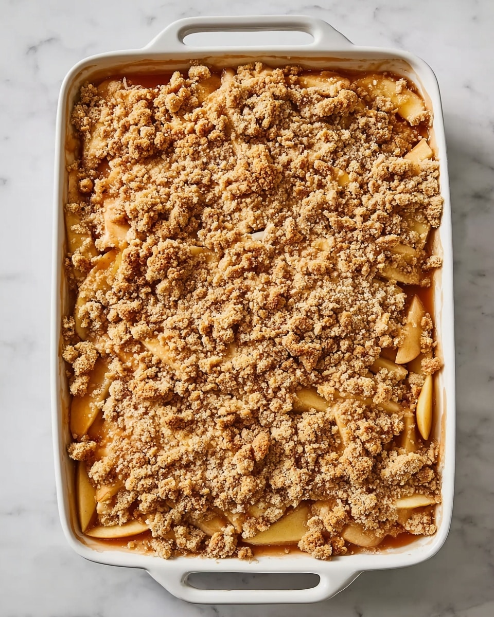 A white rectangular baking dish with handles is filled with a dessert that has three main layers visible from the top view. The bottom layer shows soft slices of light yellow fruit, likely apples, arranged evenly across the dish. Over the fruit, there is a thick layer of brown syrup or sauce, partly visible around the edges and between the crumbs. The top layer is made up of a golden-brown crumb topping, which looks crumbly and textured with small and medium-sized clusters scattered all over, covering the fruit and sauce completely. The dish rests on a white marbled surface. photo taken with an iphone --ar 4:5 --v 7
