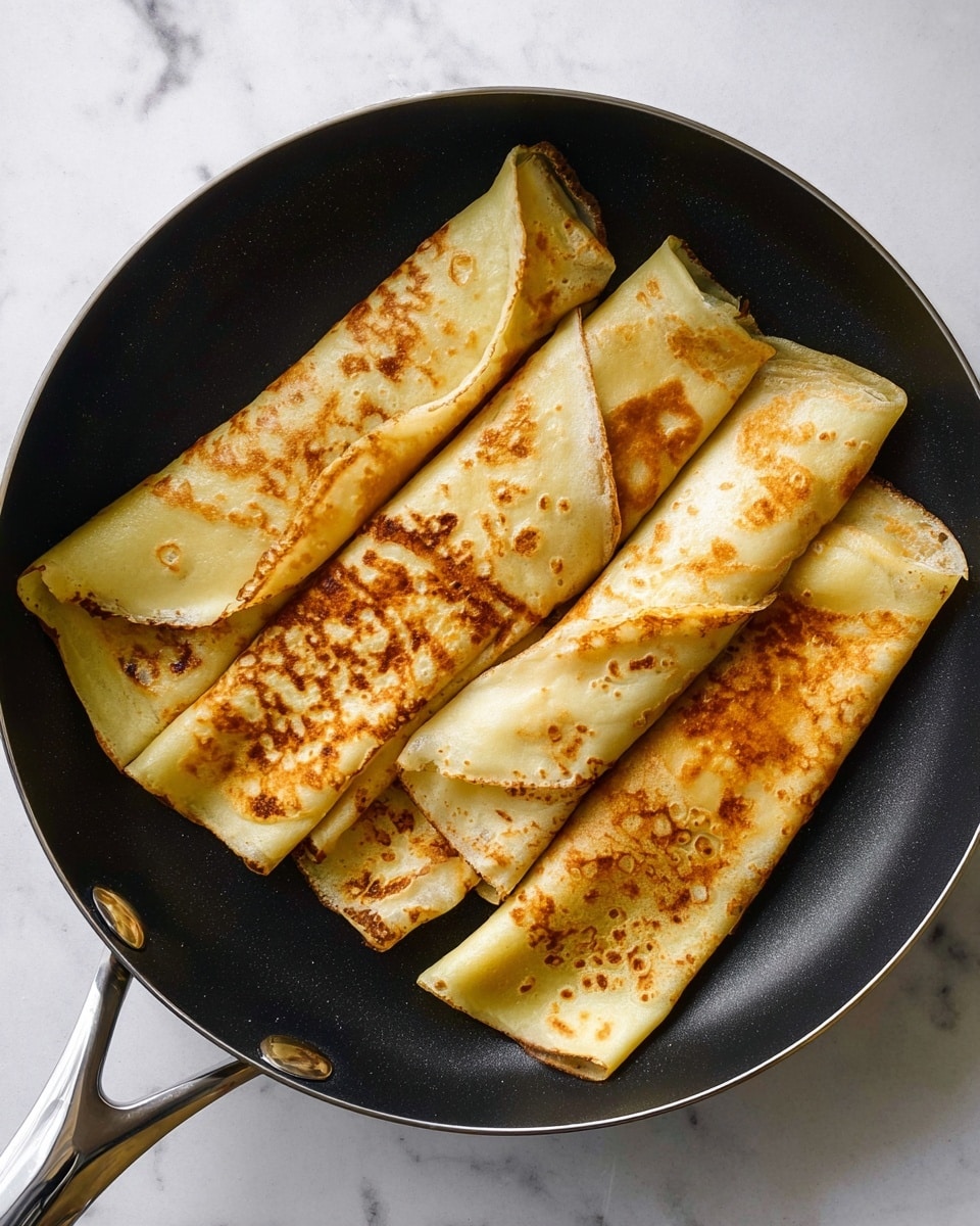 Five folded crepes are arranged in a black pan with a shiny silver handle, each crepe showing a golden-brown toasted surface with small darker spots. The crepes are folded into rectangles with visible smooth, thin layers, and they fill the pan without overlapping too much. The background is a white marbled texture. photo taken with an iphone --ar 4:5 --v 7