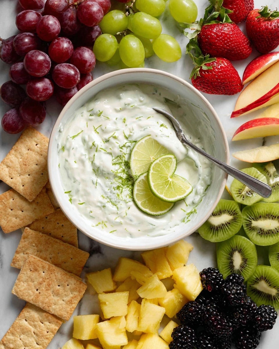A white bowl filled with creamy white yogurt mixed with green lime zest and two thin lime slices resting on top, with a silver spoon inside. Surrounding the bowl on a white marbled surface, there are fresh red grapes on top left, green grapes on top right, slices of red and green apples on the far right, halved strawberries next to the apples, green kiwi fruit slices below the apples, a cluster of blackberries below the bowl, several rectangular yellow pineapple pieces below and left of the bowl, and light brown square crackers on the left side. The dish is colorful with fresh fruit and light textures. photo taken with an iphone --ar 4:5 --v 7