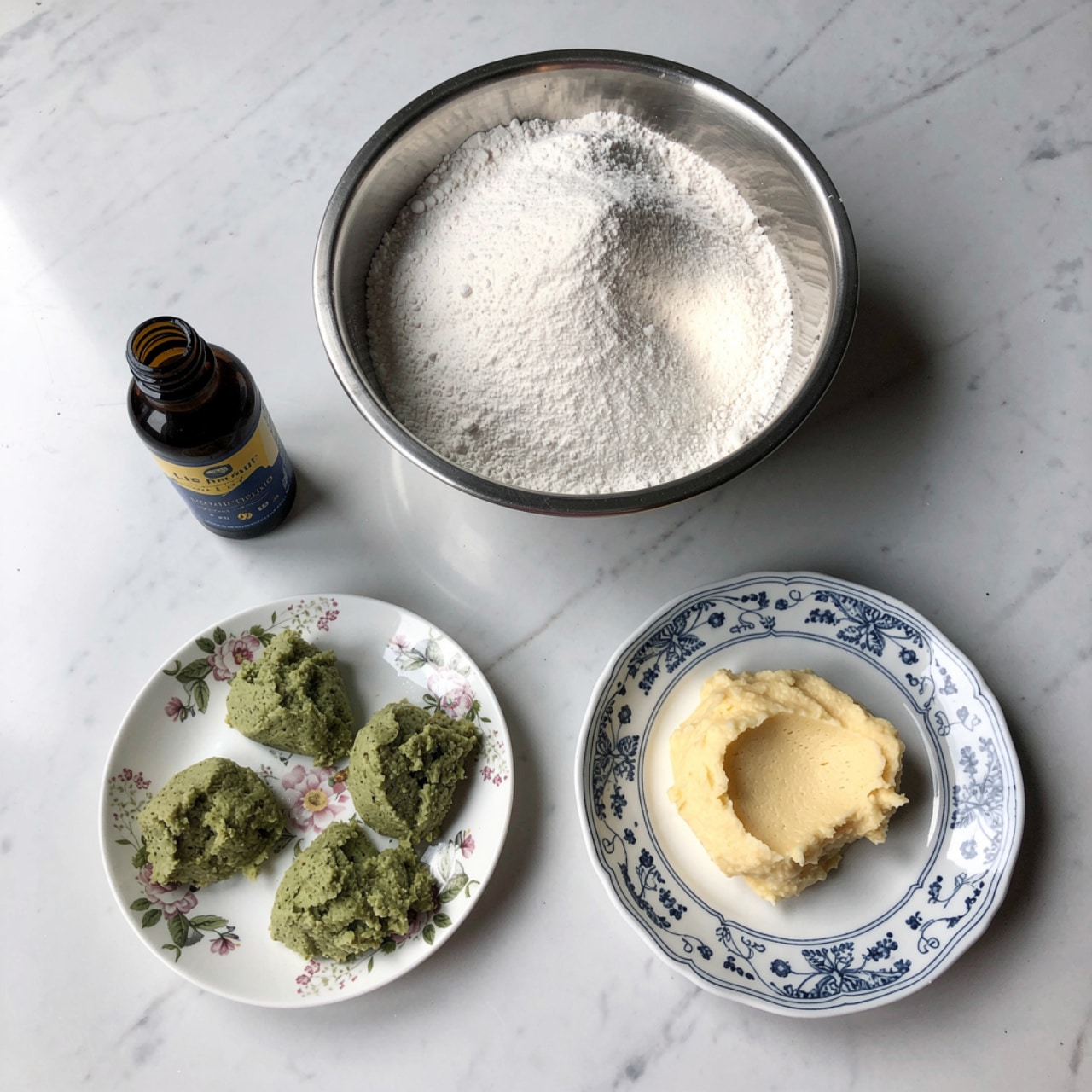 The image shows a wooden surface with four items arranged in a loose square shape. At the top right, there is a large metal bowl filled with a mound of fine white powder. At the bottom right, there is a small white plate with a blue intricate pattern holding a soft, pale yellow spread. At the bottom left, a small white dish with a floral pattern contains several small mounds of greenish paste. To the left of the metal bowl and above the green paste is a small dark brown bottle with a blue and yellow label. The background surface is changed to a white marbled texture. photo taken with an iphone --ar 4:5 --v 7