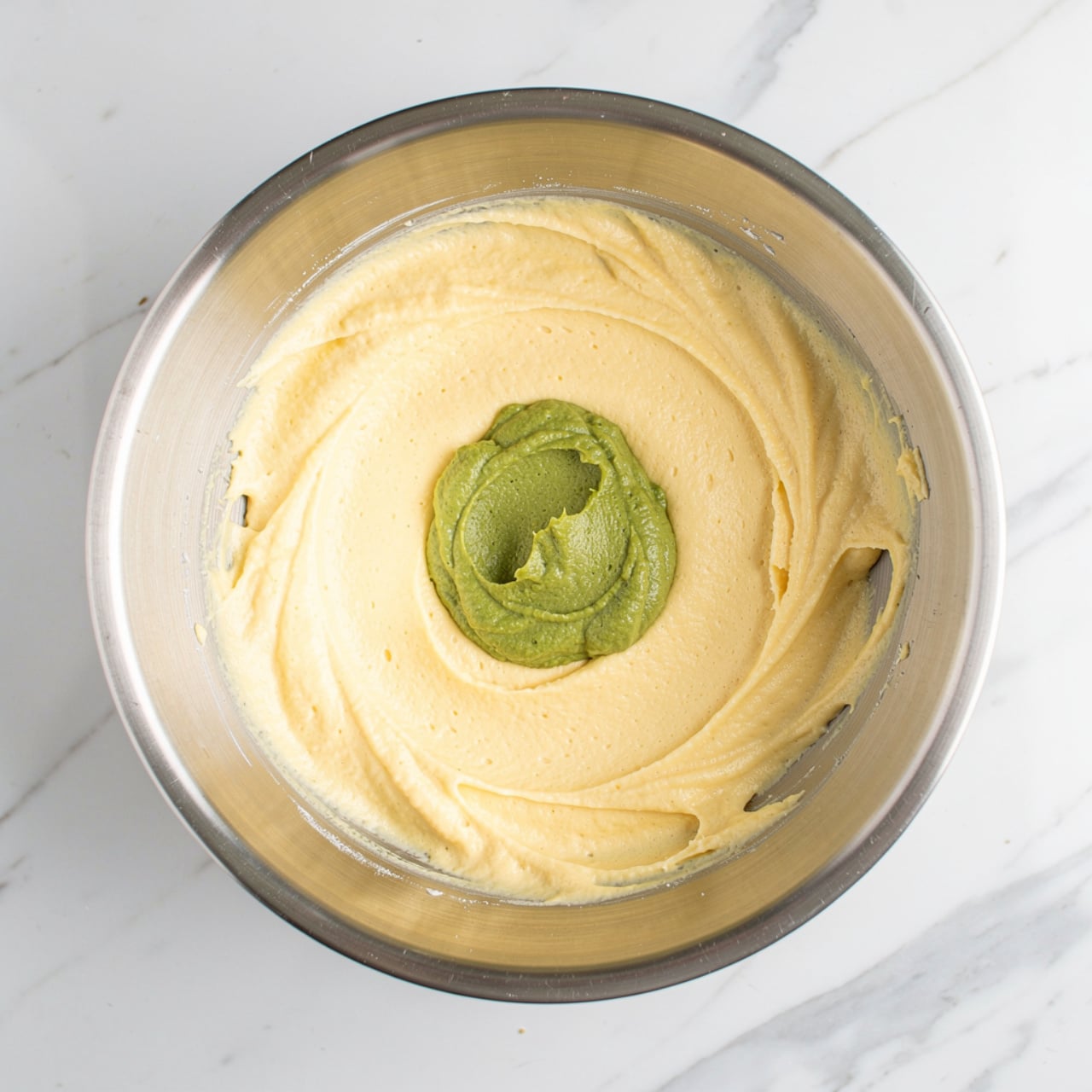 Inside a silver metal mixing bowl, there is a layer of smooth, creamy pale yellow mixture at the bottom, with a smaller dollop of thick green paste placed on top of it near the center. The bowl sits on a white marbled surface, and the image is taken from above, showing a clear view of the two textured layers inside the bowl photo taken with an iphone --ar 4:5 --v 7