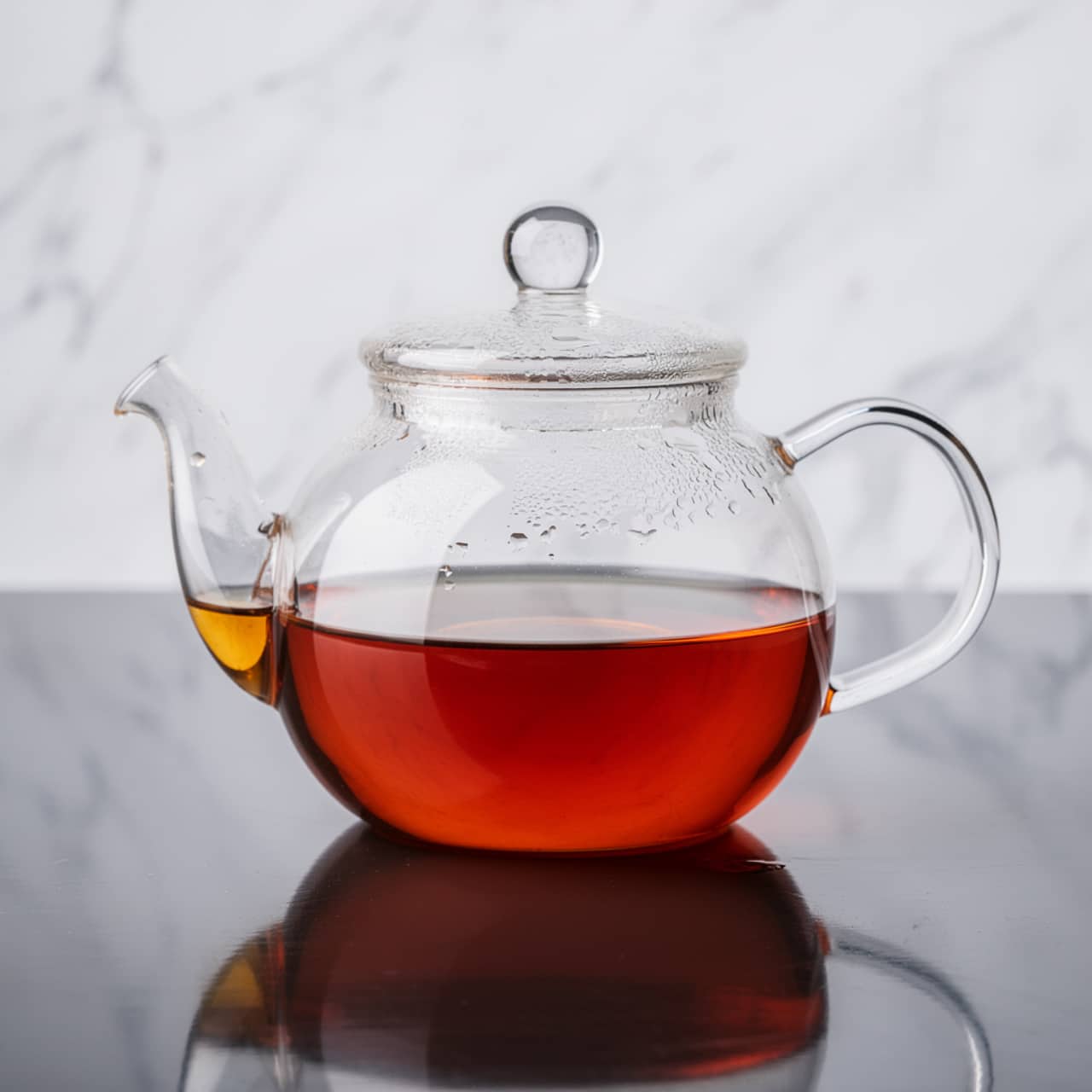 A clear glass teapot filled halfway with amber-colored tea, showing tiny droplets of steam clinging to the inside of the pot and lid. The teapot is round with a curved handle on the right and a spout on the left, sitting on a shiny dark surface that reflects its shape. The background is a soft white marbled texture, giving a clean and simple look. The photo taken with an iphone --ar 4:5 --v 7