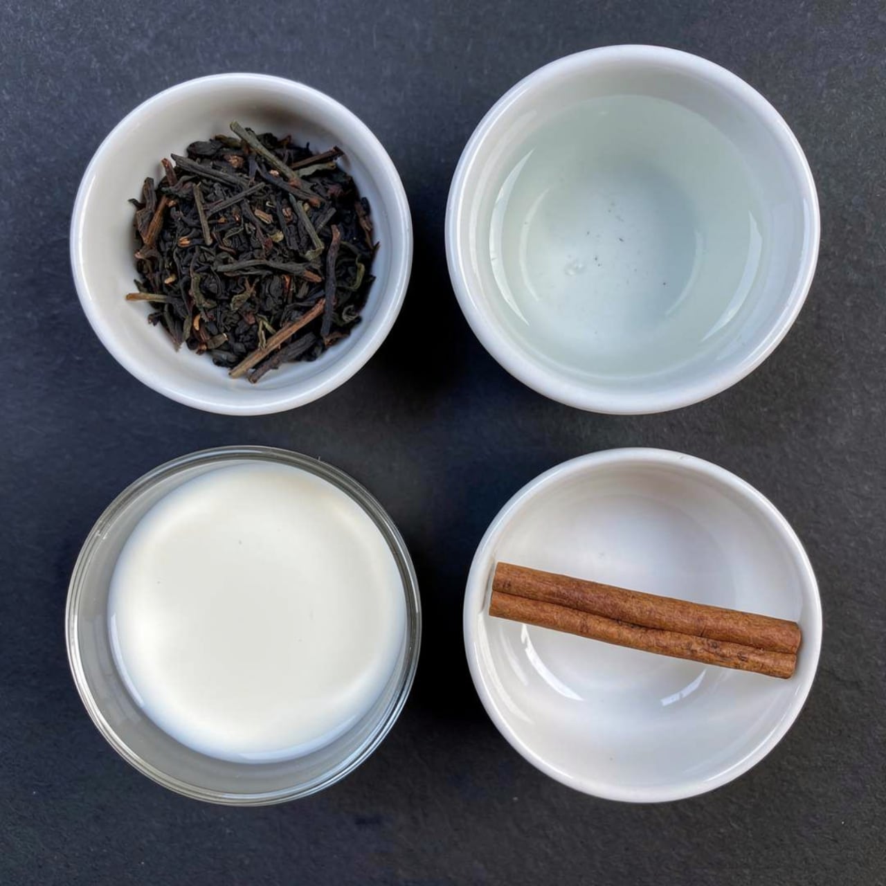 The image shows four small containers arranged on a dark surface. The top left container is a white bowl filled with loose black tea leaves with a dry, leafy texture. Next to it on the right is another white bowl holding a clear liquid, likely water, with a smooth surface reflecting light. Below the tea leaves is a clear glass cup filled with white milk, showing a creamy texture. The bottom right is a white bowl with a single cinnamon stick placed horizontally, its rough, textured brown color standing out against the simple bowl. The setting is neat and simple. Photo taken with an iphone --ar 4:5 --v 7