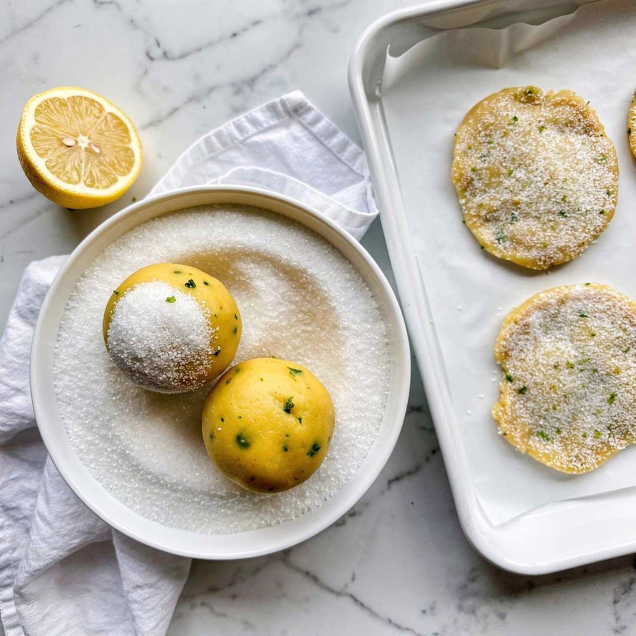 The image shows two yellow dough balls with a smooth texture and some small green specks, resting in a white bowl filled with a layer of coarse, white sugar. One dough ball is partially coated with sugar while the other is plain. To the right, a white baking tray lined with parchment paper holds three flattened dough pieces covered in white sugar, showing a rough grainy texture. There is also a half lemon with visible juicy pulp to the left on a white marbled surface. A crumpled white cloth is placed near the bowl, adding softness to the scene. Photo taken with an iphone --ar 4:5 --v 7